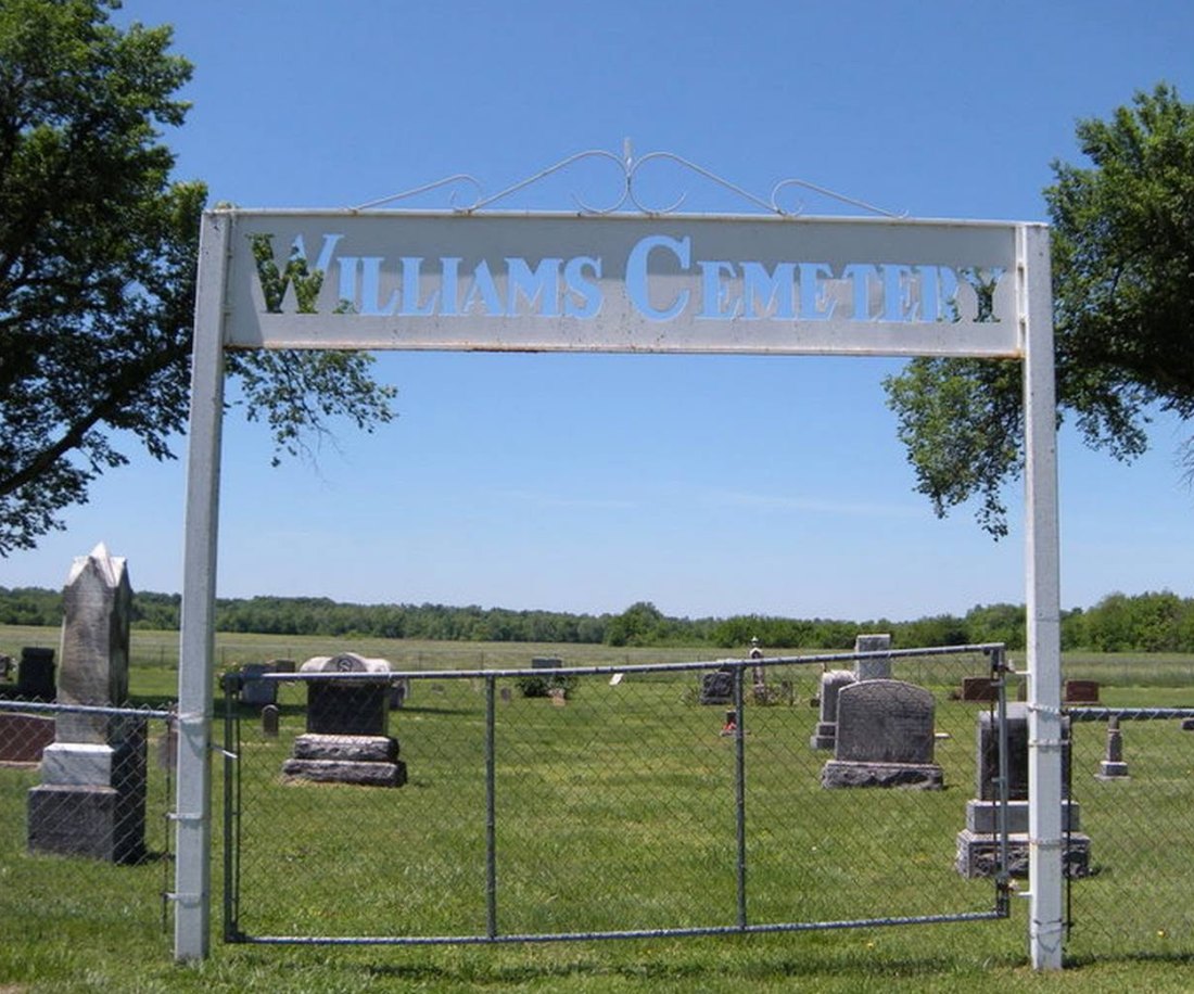 The white metal arch over the entrance to Williams Cemetery, with headstones and open country behind it.